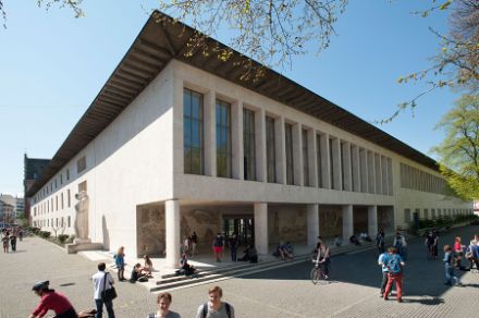 The Kollegienhaus of the University of Basel, a modern building with a minimalist design, featuring a flat roof and large entrance pillars. The facade is adorned with a colorful mural, and blue banners hang from the roof. The surrounding area is filled with lush green trees, and several people are sitting on the steps or walking by under a bright blue sky with scattered clouds.