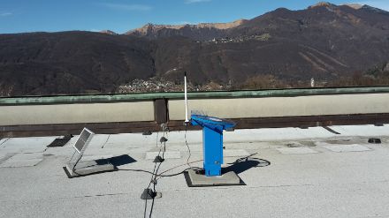 A blue, metal hail sensor with a polycarbonate measuring plate on top, next to a solar unit.