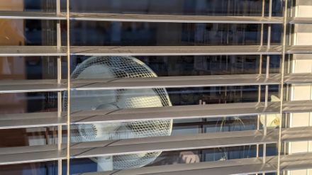  	View from outside through a window with half-closed sunblinds. There is a fan by the window inside the room.
