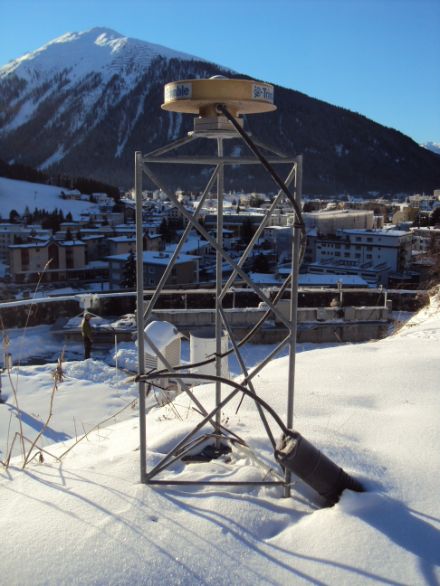 Im Vordergrund des Bildes befindet sich das GPS-Vermessungsgerät, eine metallische, auf einem Metallgerüst montierte Scheibe. Im Hintergrund ist Davos zu erkennen.