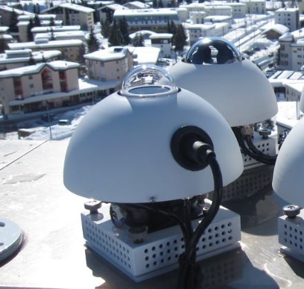 Zwei UV-Biometer, erkennbar eine kleine Glaskuppel und eine kugelförmige Abdeckung, welche die Instrumente vor Umwelteinflüssen schützt. Im Hintergrund schneebedeckte Häuser von Davos.