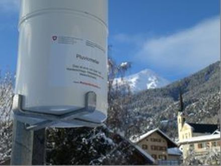 In the foreground is a pluviometer, and in the background, snow-covered forests and mountains