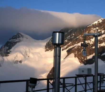 The air intakes are arranged on an open-air terrace. The instrument seen in the centre of the image consists of a tube fixed to the vertical pole and topped with a metal container across which the air is passed. Mountains and glaciers can be seen in the background.