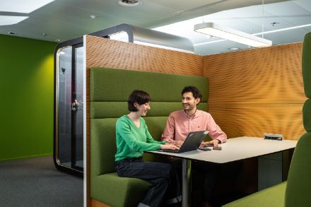 Two MeteoSwiss employees, a woman and a man, sit in a green booth with a table in the MeteoSwiss offices and work together on a laptop. There are also two cell phones on the table.